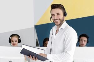 Portrait of handsome Caucasian call centre operator with headset holding documents at modern office. Male customer support manager smiling and his coworkers at open workplace photo