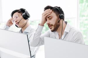 Exhausted call centre operator and his colleagues communicating with clients at open space office photo