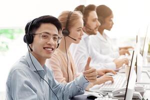 Call center worker showing thumb up gesture and his team with headphones working at open space office photo