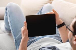 Surfing the internet. Young man with glasses and with smartwatch lies on couch at home and holds tablet with blank screen, cropped photo
