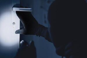 Vandalism Concept. Rear view of disguised burglar entering enclosed property, holding flash light, looking at lock photo
