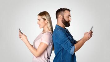 A married couple stands back to back, each engrossed in their smartphones. They appear disconnected despite their close proximity, illustrating modern relationship challenges. photo