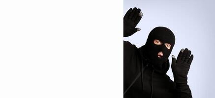 Scared thief in a balaclava and gloves raising hands up and looking out of blank sign with copyspace for your text photo