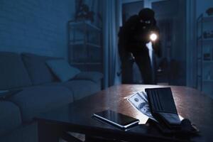 Masked thief sneaking into apartment, using flashlight, selective focus on smartphone and wallet with money lying on table photo