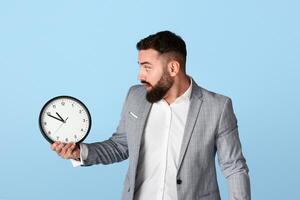 Time management problem. Scared corporate worker with analog clock missing deadline on blue background photo