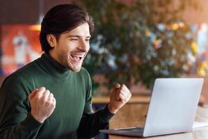 Emotional man looking at laptop, clenching fists and screaming, got good news, empty space photo