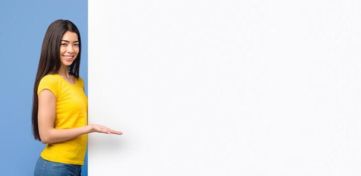 Positive Korean Lady Demonstrating Copy Space At White Advertisement Board, Pointing With Palm At Empty Place On Blank Billboard, Standing Over Purple Background And Looking At Camera photo