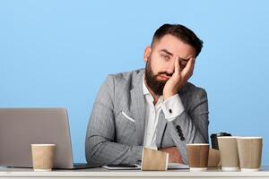 Tired businessman surrounded by empty coffee cups sitting at his desk with laptop computer against blue background photo