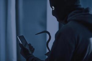 Robbery Concept. Masked villain with crowbar and smartphone preparing burglary via smart home app in the dark photo