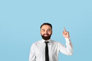 Happy millennial businessman in formalwear pointing upwards at empty space on blue studio background. Attractive bearded CEO having creative idea, gesturing eureka, experiencing aha moment photo