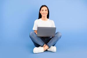 Technology. Portrait of a smiling young woman sitting with legs crossed, using laptop computer isolated over blue studio background, looking at camera. Satisfied lady studying or working with notebook photo