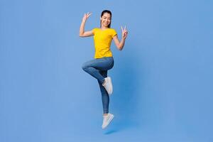 Carefree Mood. Cheerful Asian Girl Jumping In The Air And Showing Peace Gesture Over Purple Background, Full Length Shot With Free Space photo