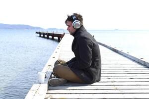 Young gloomy man listening to music sitting all alone on pier on a windy spring day. photo