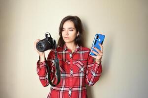 Young brunette woman photographer in red shirt choosing between camera and mobile phone on beige background. Concept of choice and difference between mobile photography and camera. Copy space photo