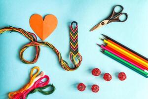 Unfinished woven friendship bracelet with skein of thread, scissors, cut heart and selected palette of crayons colors on white background photo