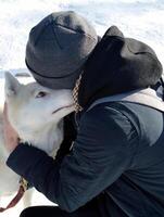 Sled siberian husky carefully and attentively sniffing the young guy in dark grey hat, who hugs the dog on a snowy background photo