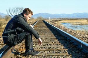 Young man in coat looking down with upset face,sitting on rails. Depressive concept loss of money, loneliness, unemployment. Copy space photo