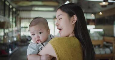Handheld zoom in shot, Young asian mother stands in a coffee shop, smiling warmly as she gently cradles a baby boy in her arms with a relaxing atmosphere during happy times video