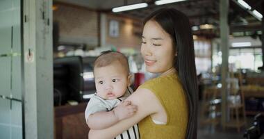 Handheld shot, Beautiful asian mother stands in a coffee shop, smiling warmly as she gently cradles a baby boy in her arms with a relaxing atmosphere during happy times video