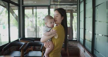 Handheld selective focus shot, Beautiful mother stands in a coffee shop, smiling warmly as she gently cradles a baby boy in her arms with a relaxing atmosphere during happy times video