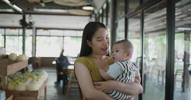 Handheld shot, Beautiful mother stands in a coffee shop, smiling warmly as she gently cradles a baby boy in her arms with a relaxing atmosphere during happy times video