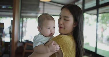 Handheld shot,Young asian mother stands in a coffee shop, smiling warmly as she gently cradles a baby boy in her arms with a relaxing atmosphere during happy times video