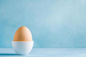 A whole egg sits in a white egg cup against a light blue background creating a minimalistic scene photo
