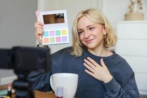 Lifestyle blogger, records in her room, has a camera on coffee table, shows eyeshadow palette to her followers, does makeup tutorial, vlogger working indoors, creates content for social media photo