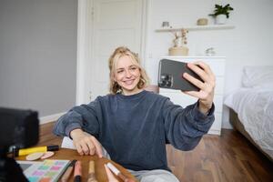 Image of young woman, vlogger taking selfie in her room, talking to her followers during online live stream, using smartphone app to chat with audience, smiling and looking happy photo