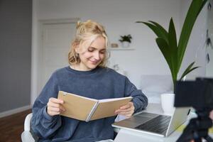 Portrait of young woman using digital camera and laptop, studying, connect to online live stream, records for vlog, creating content, holding notebook, sitting in room photo