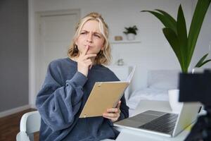 Portrait of young woman, student sitting in room, studying from home, remote education concept, holding notebook and thinking, looking aside with thoughtful face, concentrating on assignment photo