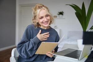 Portrait of smiling, beautiful young blond woman, student working on assignment from home, online learning in her bedroom, talking to camera, chatting, holding notebook photo