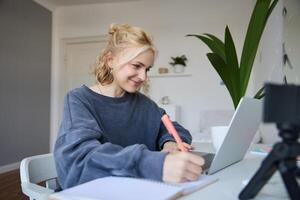 Portrait of smiling blond woman writing in notebook, making notes, recording content for social medial on digital camera, looking at laptop, working or studying photo