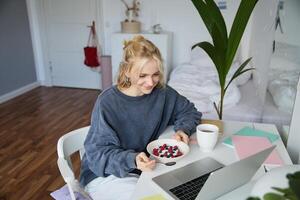 Portrait of smiling blond young woman, eating in front of laptop, watching s online while having breakfast, enjoying dessert, sitting in bedroom photo