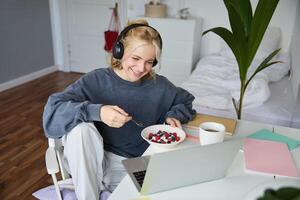 Portrait of smiling young blond woman in headphones, sitting in room, watching movie on laptop, eating breakfast and drinking tea, having lunch in front of computer screen photo