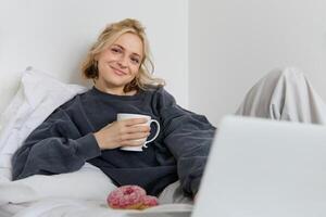 Portrait of happy woman enjoying day-off at home, lying in bed, watching tv show on laptop, looking at screen and smiling, drinking tea and eating in bedroom photo