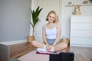 Portrait of young fitness instructor, vlogger showing exercises on camera, recording herself, sitting on mat with laptop, doing workout, explaining yoga movements to followers photo