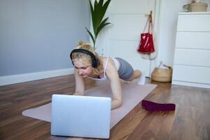 Portrait of young athletic woman in wireless headphones, standing in plank, following online workout , using laptop to join training session on the internet photo
