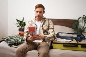 Portrait of young man, tourist is packing his luggage, looking at tour guide, travel map, sitting on bed with his clothes in a suitcase photo