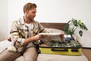 Tourism and travelling concept. Portrait of handsome young man going on holiday, packing his suitcase, checking luggage, sitting on bed and looking at packed things photo