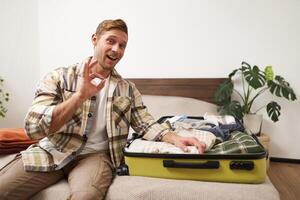 Enthusiastic young man, tourist gets ready for holiday, sits on bed with suitcase and shows okay, approval sign, recommends good quality luggage photo