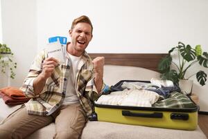 Portrait of happy, excited young man, tourist holds his passport and plane tickets, packs clothes in suitcase, gets ready for summer holiday, goes on vacation and feels excitement photo
