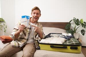Portrait of handsome young man, traveller shows his passport and two tickets, packing suitcase with clothes, going on vacation photo