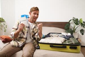 Portrait of cheerful guy sits near his luggage, packs suitcase, holds two plane tickets and passport, goes on holiday, books vacation stay photo