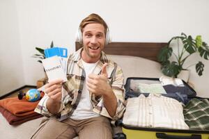 Portrait of smiling man, tourist shows two tickets and passport, goes on holiday, traveller makes thumb up in approval, like travel agency photo