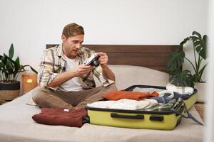 Portrait of young man looking at his digital camera screen, checking photo album on gadget, unpacking suitcase after holiday, travelling with luggage, sitting in hostel on a bed