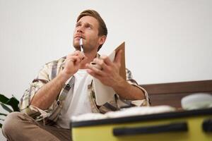 Portrait of tourist packing suitcase, going through check-list, writing down items to take with him in luggage, making notes in notebook, sitting on bed photo