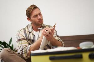 Portrait of tourist packing suitcase, going through check-list, writing down items to take with him in luggage, making notes in notebook, sitting on bed photo