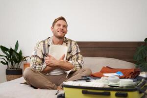 Portrait of thoughtful young man, tourist planning his vacation, sitting with open suitcase on bed, making notes, writing to do check list in notebook photo