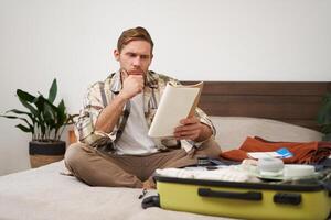 Portrait of man with suitcase, sits in room and looking serious at his notes, packing for holiday, checking list of items in notebook photo